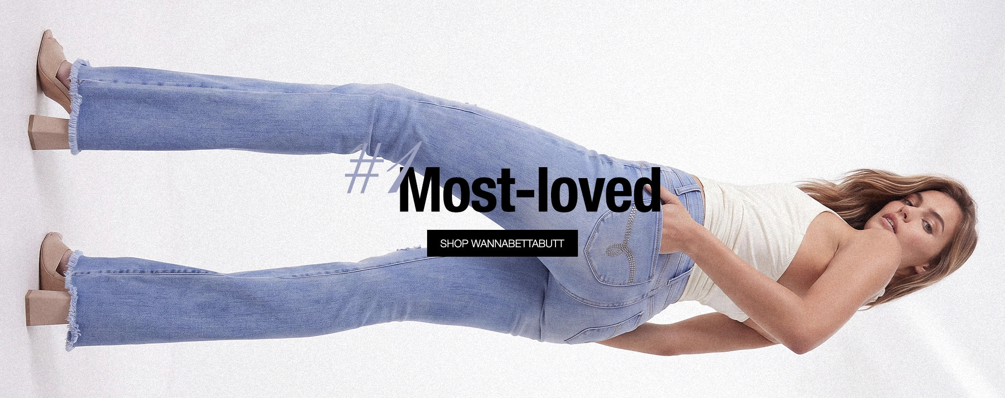 A woman lying on her side against a white studio background, wearing light-wash blue jeans and a white sleeveless top. Large overlaid text reads '#Most-loved' with a smaller 'SHOP WANNAWITNESSOUT' below.