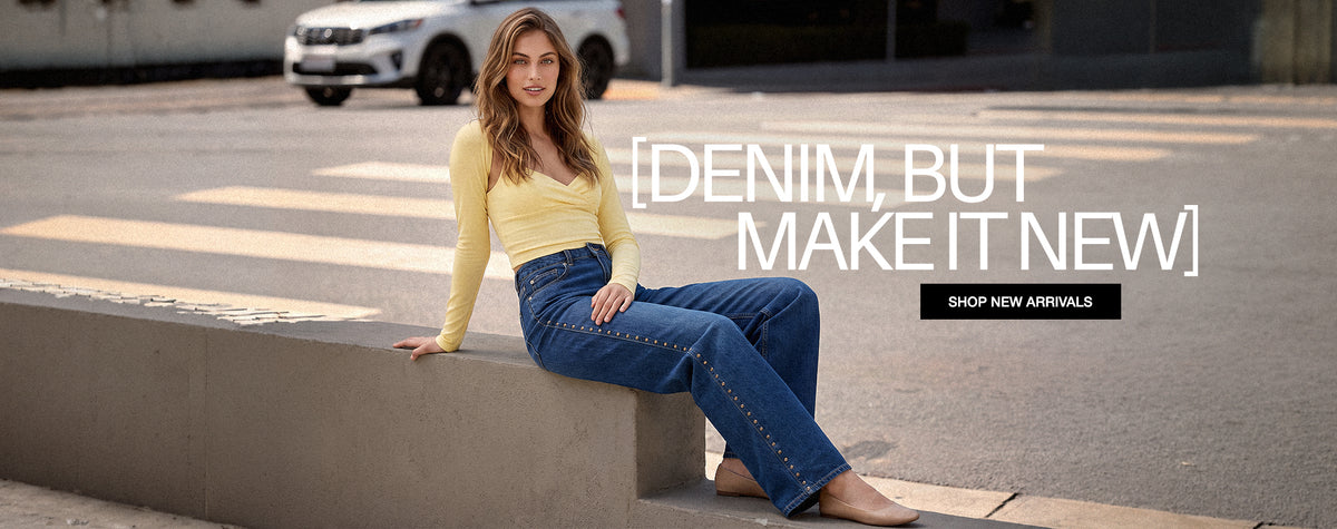 A woman sits on a concrete curb in an outdoor street setting, wearing a yellow long-sleeve top and blue denim jeans. Large overlaid text reads '[DENIM, BUT MAKE IT NEW]' with a smaller 'SHOP NEW ARRIVALS' button.