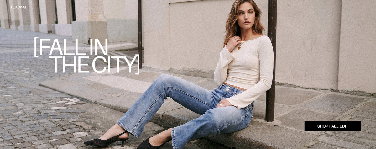 A woman sits on a stone ledge outdoors wearing a cream ribbed long-sleeve top, light blue flared jeans, and black heels. Large white text reads '[FALL IN THE CITY]' on the left, with a small black 'SHOP FALL EDIT' box in the bottom right.