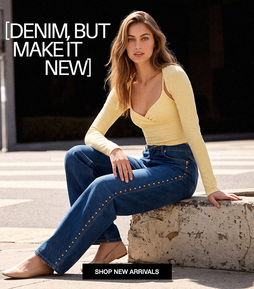 A woman sits on a stone curb outdoors wearing blue denim jeans with gold-tone stud detailing along the outer seams, paired with a pale yellow long-sleeve wrap top. Large text at the top reads '[DENIM, BUT MAKE IT NEW]' and a black banner at the bottom says 'SHOP NEW ARRIVALS'.