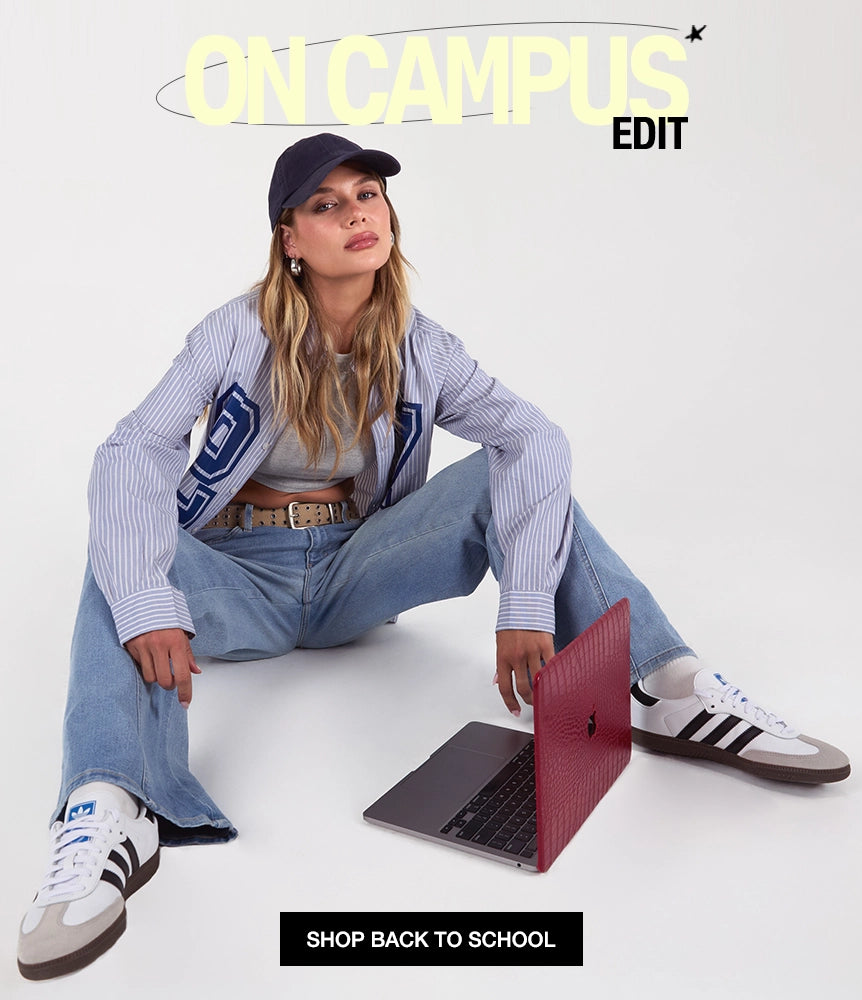 A woman sits cross-legged on a white studio floor wearing a navy cap, a light blue and white striped button-up shirt with large navy lettering, light-wash jeans, and white sneakers with black stripes. A red textured laptop skin covers a laptop in front of her.