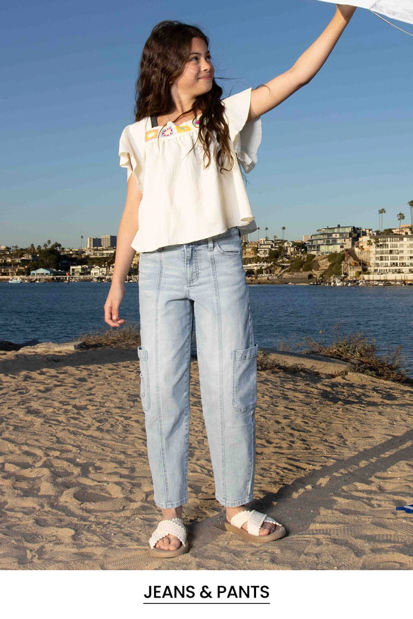 Woman standing on a beach wearing light blue jeans and a white top, with a scenic background.